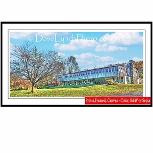 May include: A vintage-style photograph of a blue building with a large porch and a grassy lawn. The building has a stone facade and a wooden roof. There are trees in the foreground and a blue sky with white clouds in the background.