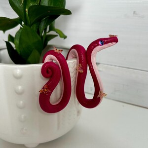 Peut inclure: Une figurine de serpent rose et blanc avec des accents de papillon. Le serpent est enroulé autour d'un pot blanc.