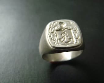 May include: A silver signet ring with a square face featuring an engraved crest. The crest is detailed with intricate floral designs and a central symbol.