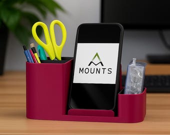 May include: A burgundy desk organizer with a smartphone displaying the word "MOUNTS" and a logo. The organizer holds pens, yellow scissors, and a clear container. A computer and plant are in the background.
