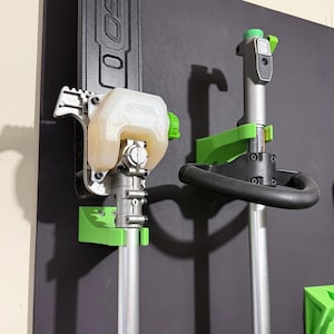 Può includere: Due supporti a parete verdi per un decespugliatore. I supporti sono in plastica e hanno una combinazione di colori verde e nero. Il decespugliatore è uno strumento a benzina con un serbatoio del carburante trasparente.