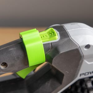 May include: Close-up of a green plastic strap securing a black and gray power tool. The tool has a green button with an arrow symbol. The word "POWER" is visible on the tool's body.