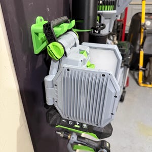 May include: A green and gray wall mount for power tools. The mount has a black handle and a green and gray power tool is attached to it. The power tool has a gray body and a green handle. The power tool has a power button and a plus and minus sign on it.