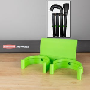 May include: A lime green vacuum cleaner accessory holder with two curved cradles and a back panel. A framed image shows black vacuum attachments. The background features a wooden surface and a gray wall.