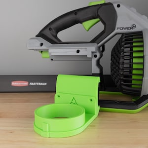 Peut inclure: Un souffleur de feuilles sans fil avec une poignée noire et des accents verts. Un support vert vif est au premier plan. Le souffleur est gris et noir avec le mot "POWER" imprimé sur le côté. Le support est conçu pour maintenir le souffleur.