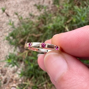 May include: A gold ring set with small, bright red gemstones. The ring has a band design, with the red stones arranged in a line. The ring is held up against a blurred green backdrop.
