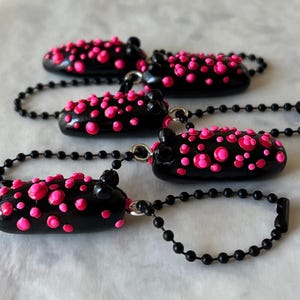 May include: Four black keychains with pink polka dots. Each keychain has a black ball chain and a silver-coloured clasp. The keychains are oval-shaped and have a glossy finish.