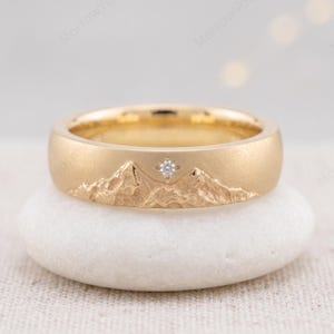 May include: A gold band ring featuring a textured mountain range relief across the outer surface. A small, clear, round-cut gemstone is set into the metal above the mountain peaks, designed in a four-pointed star shape setting.