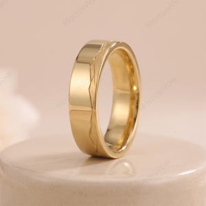 May include: A polished gold-tone band ring featuring a smooth, flat surface on one side and a series of angular, geometric etched lines creating a subtle mountain-like pattern along the outer edge of the metal band.