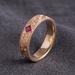 May include: A rose gold band ring featuring an intricate engraved scrollwork pattern along the outer surface. The ring is accented with a single square-cut red ruby gemstone set within a diamond-shaped frame, bordered by a delicate beaded edge.