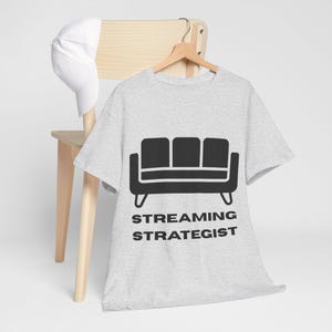 以下が含まれることがあります： 黒いソファのグラフィックと「STREAMING STRATEGIST」の文字がプリントされた、明るい灰色のTシャツ。白い野球帽が、Tシャツの後ろにある木製の椅子の上に置かれています。