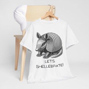 Op de afbeelding: Wit T-shirt met een zwart-witte illustratie van een gordeldier. De tekst "Let's Shellebrate!" is onder het gordeldier gedrukt.