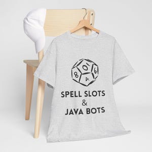 May include: A light grey t-shirt featuring a black die graphic and the text "SPELL SLOTS & JAVA BOTS." A white baseball cap is on a wooden chair in the background.