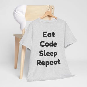 Könnte beinhalten: Ein hellgraues T-Shirt mit den Worten "Eat Code Sleep Repeat" in fetten schwarzen Buchstaben. Das Shirt hängt an einem Holzkleiderbügel. Eine weiße Baseballkappe liegt auf der Rückseite eines Holzstuhls.