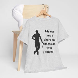 May include: A light gray t-shirt with a black silhouette of a golfer and the text "My cat and I share an obsession with birdies." A white baseball cap hangs on a wooden chair.