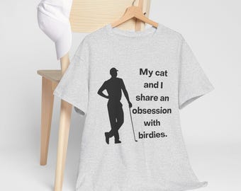 Peut inclure: T-shirt gris clair avec une silhouette noire d'un golfeur et le texte "My cat and I share an obsession with birdies." Une casquette de baseball blanche est suspendue à une chaise en bois.