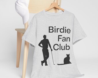 Peut inclure: Un t-shirt gris clair avec les mots "Birdie Fan Club" en noir. Le t-shirt présente une silhouette noire d'un golfeur et d'un chat. Une casquette de baseball blanche repose sur une chaise en bois.