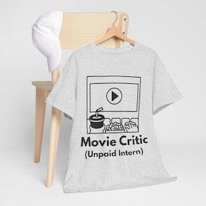 以下が含まれることがあります： 映画のスクリーン、飲み物、ポップコーン、映画館の座席の黒いグラフィックが描かれた、明るい灰色のTシャツ。「Movie Critic (Unpaid Intern)」の文字が下にあります。白い野球帽が木製の椅子の上に置かれています。