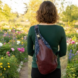 Op de afbeelding: Een bruine leren sling bag met een geometrisch patroon stoffen paneel en zwarte band. De tas wordt over de schouder gedragen. De achtergrond is een tuin met kleurrijke bloemen.