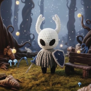 May include: A handmade crochet figure of a character with a white head, large black eyes, and two curved horns. It wears a gray tunic, blue cape, and holds a sword and shield. The figure stands on a grassy surface with a forest backdrop.