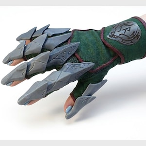以下が含まれることがあります： 緑とマルーンの指なし手袋に、灰色の角張った爪が付いています。手袋には手首に装飾的なエンブレムが付いています。爪は、指の鋭い石のような延長のように見えるように設計されています。