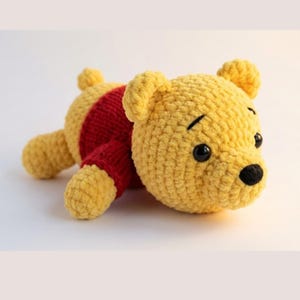 May include: A handmade yellow crochet bear with a red shirt. The bear has black eyes and a black nose. The plush toy is lying down on a white surface. The bear's ears are rounded and the body is soft and textured.