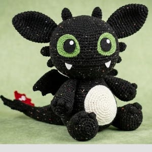 Puede incluir: Un dragón negro de ganchillo hecho a mano con grandes ojos verdes, dientes blancos y una barriga color crema. El dragón tiene alas negras y una cola roja y blanca. El peluche está sentado sobre una superficie verde.
