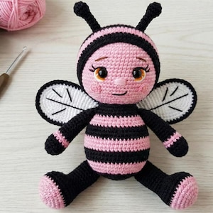 May include: A handmade crochet bee doll. It features a pink and black striped body, black limbs, and white wings. The bee has a smiling face with brown eyes and antennae. The doll is sitting on a light-coloured surface.