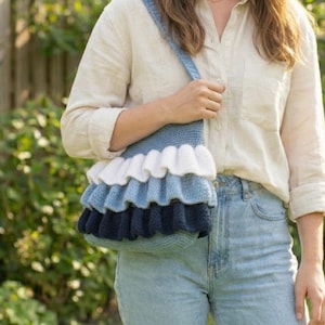 May include: A crochet shoulder bag in shades of blue, white, and navy, featuring a layered, ruffled design. The bag has a long strap and is being held. The bag is made of soft yarn and is a handmade item.