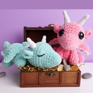 May include: Two crocheted dragon toys, one light blue and one pink, rest in a wooden treasure chest. The dragons have white horns and large black eyes. The chest contains gold coins and is set against a purple backdrop.