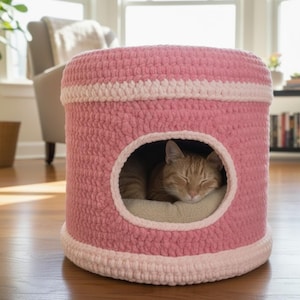 May include: A pink and white crochet cat bed with a round opening. An orange tabby cat is sleeping inside on a beige cushion. The cat bed is cylindrical and sits on a hardwood floor.