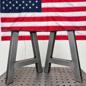 May include: Two A-frame table legs constructed from raw steel with a visible metallic gray finish. Each leg features a flat mounting plate at the top and a horizontal support bar connecting the two angled vertical posts, showing welded joints.