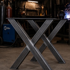 May include: A pair of industrial-style table legs in a dark gray, metallic finish. The legs are constructed in an X-shape, with a sturdy, geometric design. The legs are designed to support a tabletop. The background shows a workshop setting.