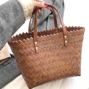 May include: A brown woven basket bag with leather handles and gold-colored rivets. The bag has a rectangular shape with a zig-zag top edge. The bag is held by a person wearing a gray blazer.