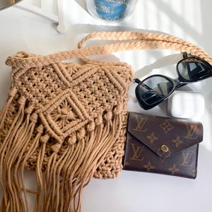 May include: A beige macrame crossbody bag with a long braided strap, fringe detail, and geometric patterns. The bag is next to black sunglasses, a white wireless earbud case, and a brown wallet with a logo.