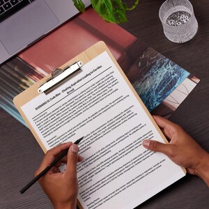 May include: A wooden clipboard holds a medical document detailing a Code Blue scenario involving choking and cardiac arrest. A black pen lies on the document. A person's hands are visible. A laptop and plant are in the background. The document is in English.