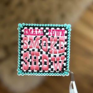 Puede incluir: Un parche bordado rosa y blanco con un fondo a cuadros. El parche dice "Small Town Smoke Show" en una fuente rosa audaz.