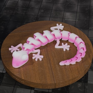 May include: A pink and white articulated lizard toy is displayed on a round wooden surface. The lizard has a segmented body and four legs with detailed feet. The toy's head and body segments transition from white to pink.