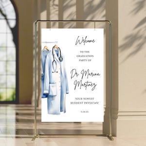 Peut inclure: Une pancarte blanche avec une illustration aquarelle d'une blouse de m&eacute;decin et d'un st&eacute;thoscope accroch&eacute;s &agrave; un cintre. Le texte indique "Bienvenue &agrave; la f&ecirc;te de graduation du Dr Marina Martinez Votre nouveau m&eacute;decin r&eacute;sident ! 5.24.25"