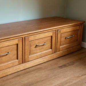 May include: A wooden storage bench with three drawers. The bench is made of light brown wood and has silver-colored handles. The bench is placed against a light green wall and sits on a wooden floor.