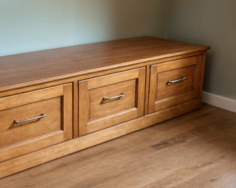 May include: A wooden storage bench with three drawers. The bench is made of light brown wood and has silver-colored handles. The bench is placed against a light green wall and sits on a wooden floor.