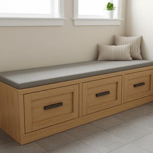 May include: A wooden storage bench with three drawers, featuring a grey cushioned top and black handles. Two striped cushions rest on the bench. The bench is made of light-coloured wood and is placed against a neutral-coloured wall.