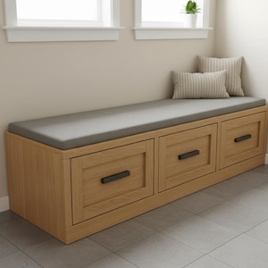 Puede incluir: Un banco de almacenamiento de madera con tres cajones y un cojín gris. El banco está hecho de madera clara y tiene tiradores negros. Dos almohadas a rayas descansan en el banco cerca de una ventana.