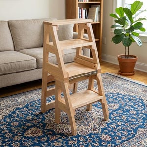 Op de afbeelding: Een lichtgekleurde houten opstap met drie treden. De opstap is gemaakt van lichtgekleurd hout en heeft een opvouwbaar ontwerp. De opstap staat op een blauw patroonkleed.