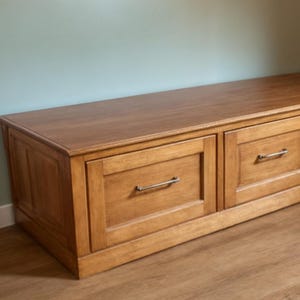 May include: A wooden storage bench with two drawers and a light brown finish. The drawers have silver-colored handles. The bench is placed against a light blue wall and sits on a wooden floor.