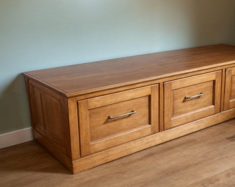 May include: A wooden storage bench with two drawers and a light brown finish. The drawers have silver-colored handles. The bench is placed against a light blue wall and sits on a wooden floor.