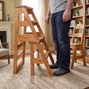 Op de afbeelding: Een houten opstapje met een ladderontwerp. Het opstapje is gemaakt van lichtbruin hout en heeft drie treden. Een persoon staat naast het opstapje. Een houten stoel en een boekenkast staan op de achtergrond.