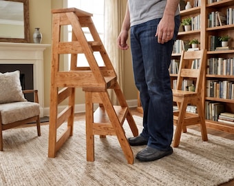 May include: A wooden step stool with a ladder design. The step stool is made of light brown wood and has three steps. A person is standing next to the step stool. A wooden chair and a bookshelf are in the background.