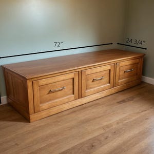 May include: A wooden storage bench with three drawers and brushed metal handles. The bench is 72 inches long, 18 inches high, and 24.75 inches deep. The bench is made of light brown wood and sits on a wooden floor.