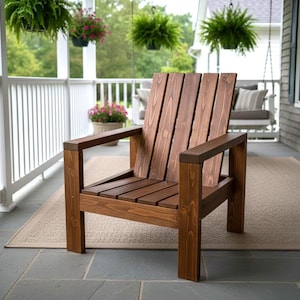 May include: A wooden garden chair with a slatted back and armrests. The chair is stained a warm brown colour and is placed on a neutral-toned rug on a porch. The chair is designed for outdoor seating.